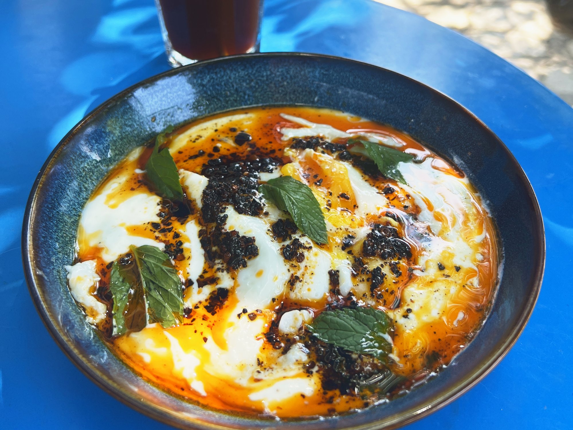 A bowl of çılbır — poached eggs on garlic yogurt with chili butter and mint — next to an iced coffee on a blue café table in Morocco