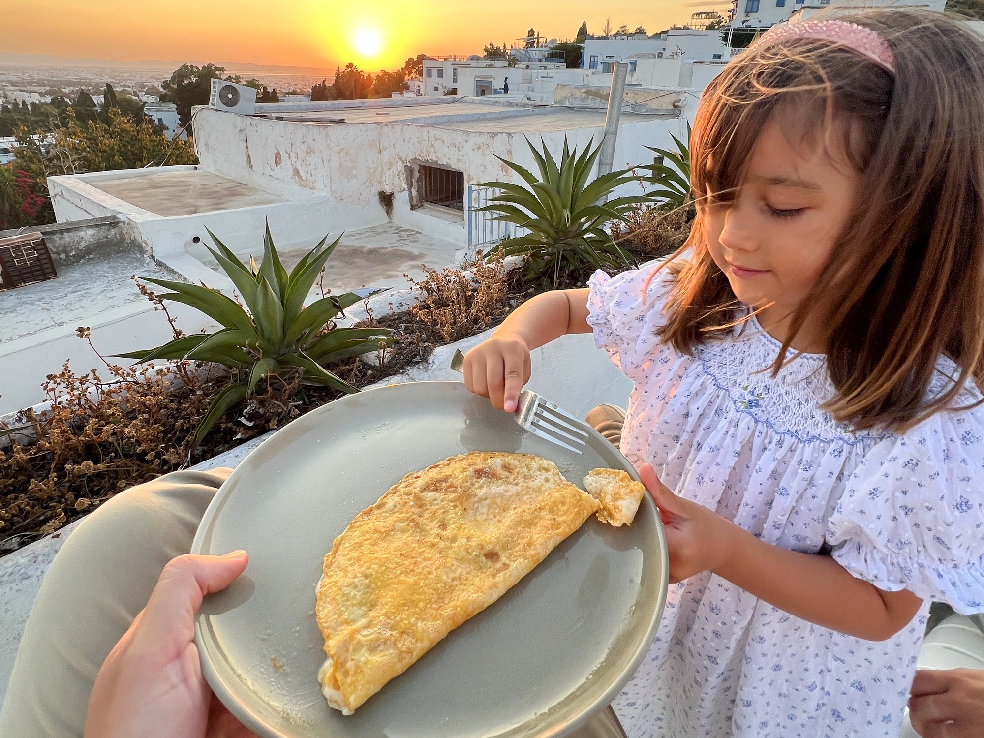 A young girl reaching with a fork to take the first bite of an omelette on a plate, on a whitewashed rooftop at sunrise in Tunisia