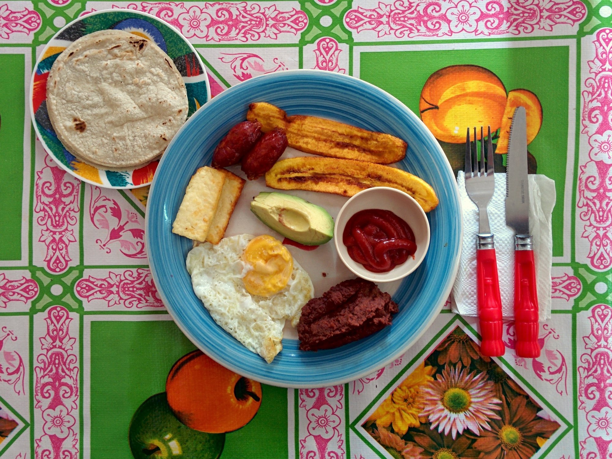 A traditional Honduran breakfast in Siguatepeque: fried egg, fried plantains, chorizo, refried beans, avocado, fried cheese and corn tortillas on a colorful oilcloth
